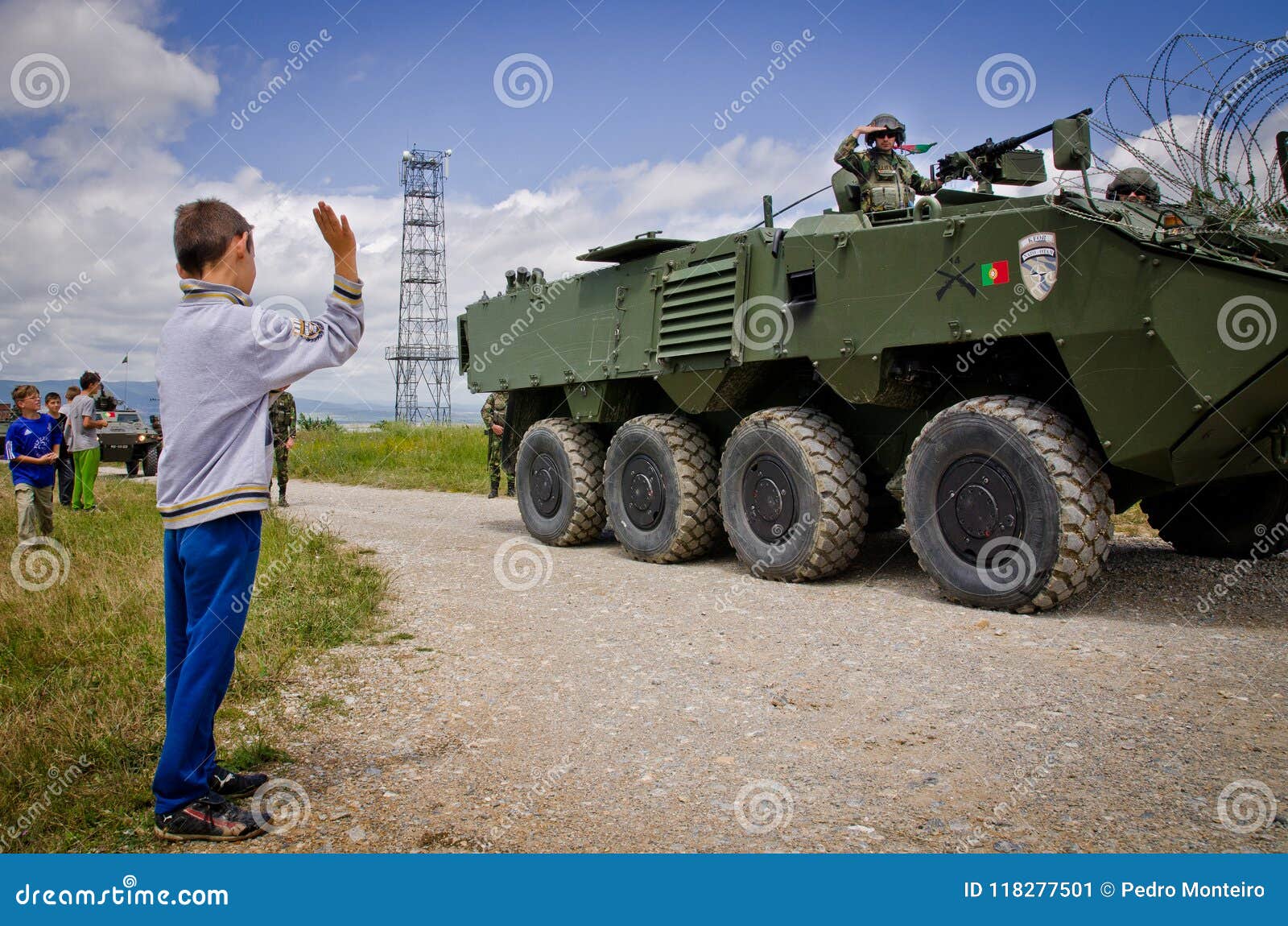 The image shows a young boy waving at a line of passing tanks. The tanks are are Portuguese under NATO command in Kosove